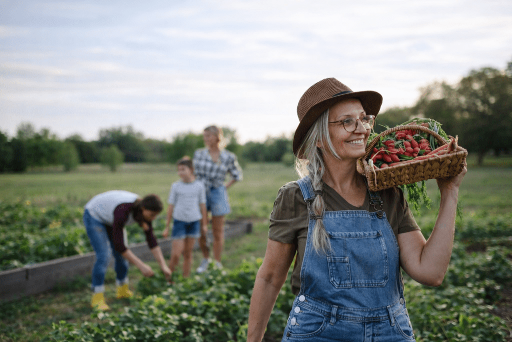 Community gardener with harvest basket An older woman smiling while carrying a basket of freshly harvested vegetables in a community garden, with volunteers working together in the background.