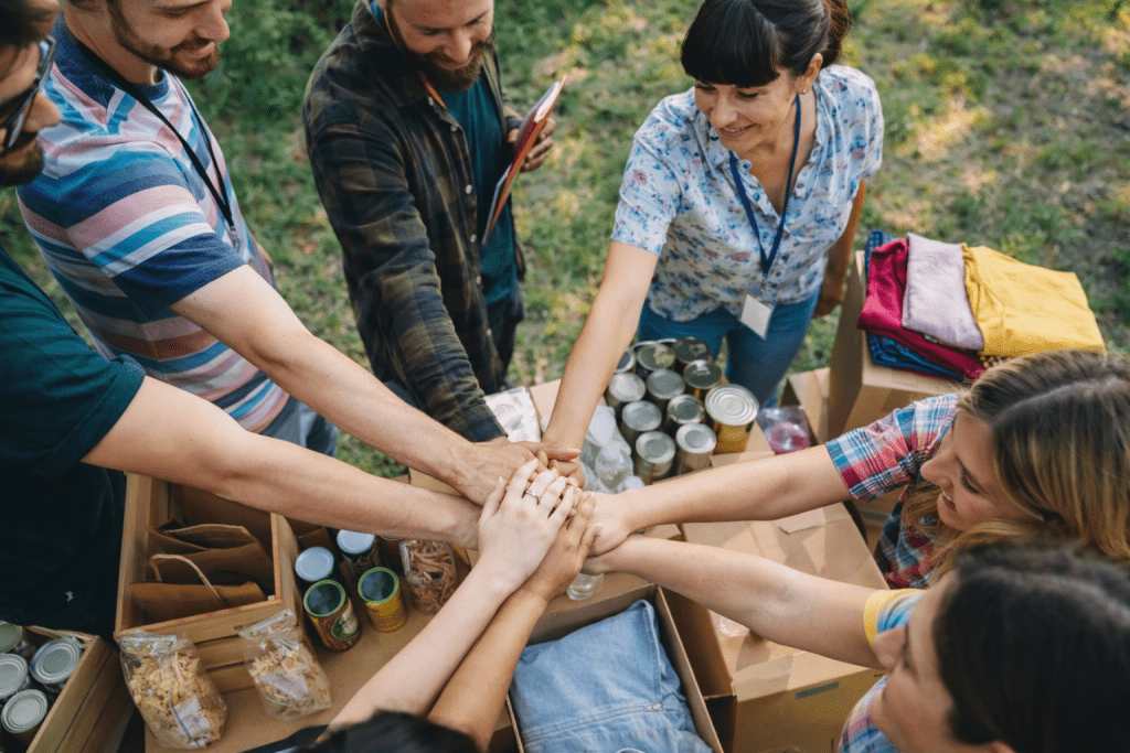 Hands Together, Impact Ahead A group of volunteers standing in a circle with their hands stacked together over boxes of donated items outdoors.